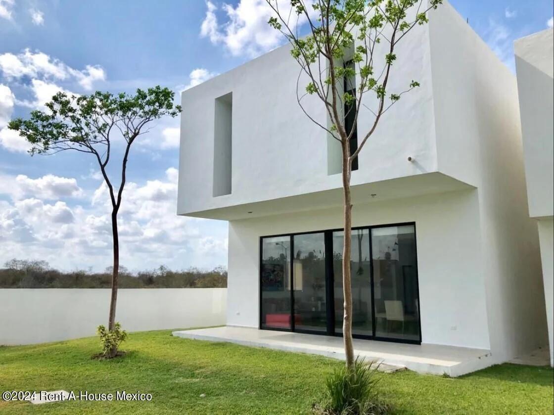 Casa moderna de dos pisos en venta o renta en la zona de Mérida, México. Presenta un diseño minimalista en blanco con grandes puertas corredizas de vidrio y un jardín verde. 