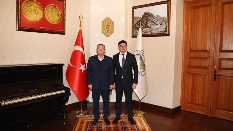 Kars Başkonsolosu ve Vali, bir odada yan yana duruyor, arka planda Türk bayrağı ve konsolosluk amblemi var.
