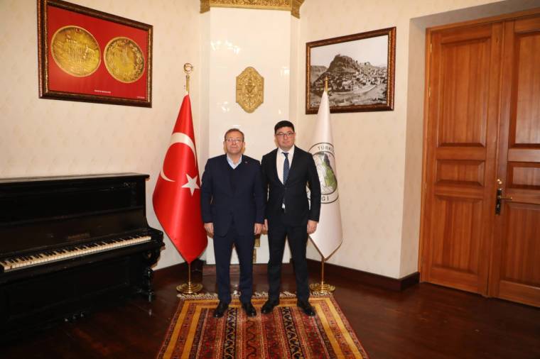 Kars Başkonsolosu ve Vali, bir odada yan yana duruyor, arka planda Türk bayrağı ve konsolosluk amblemi var.