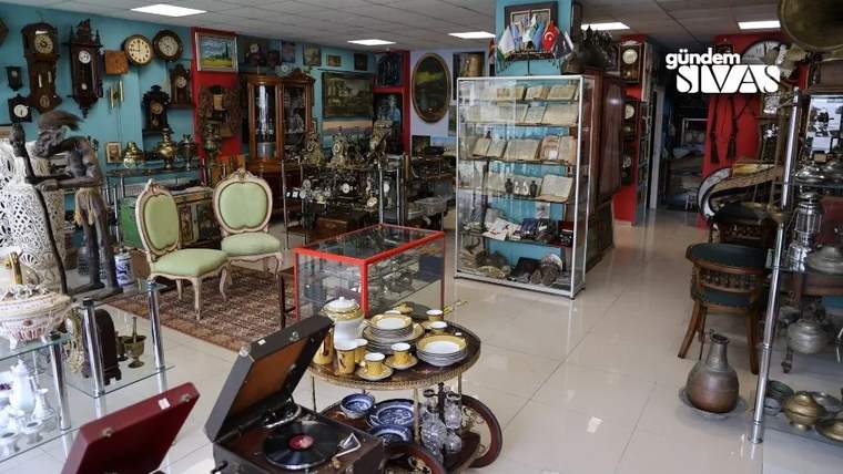 Duvar ve vitrinlerde saatler, tablolar ve çeşitli antika eşyalar; ortada tabak ve fincanların bulunduğu masalar