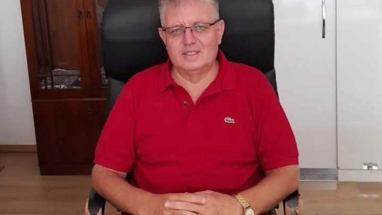 Gözlüklü bir erkek, kırmızı polo tişörtle ofiste masanın önünde ellerini birleştirerek oturuyor.