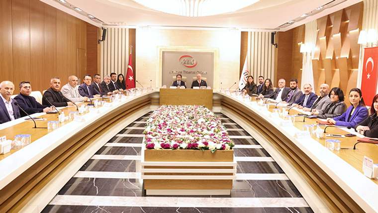 Uzun toplantı masası çevresinde oturan kişiler, ortadaki çiçek düzenlemesi ve Türk bayrağıyla birlikte görülüyor.