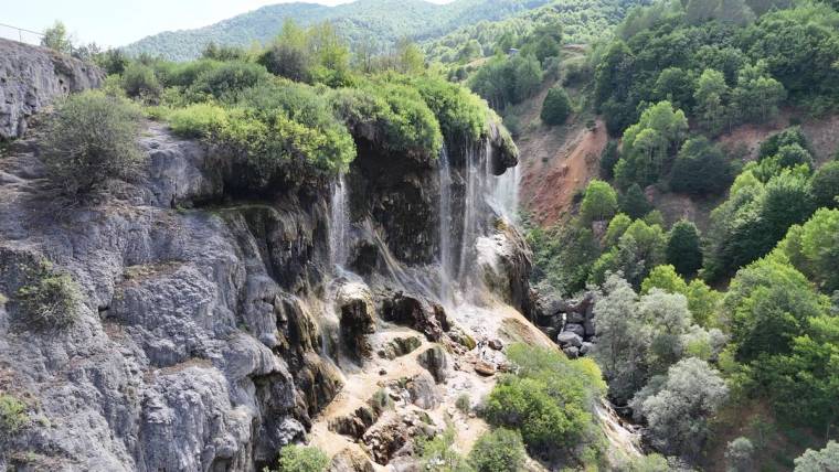 Kayalık yamaçtan ince şelaleler akıyor; çevresinde yeşil ağaçlar ve çalılıklarla kaplı tepeler görülüyor.