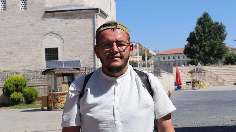 Başında takke, gözlük ve sırt çantası bulunan bir kişi, taş bir yapının önünde duruyor.