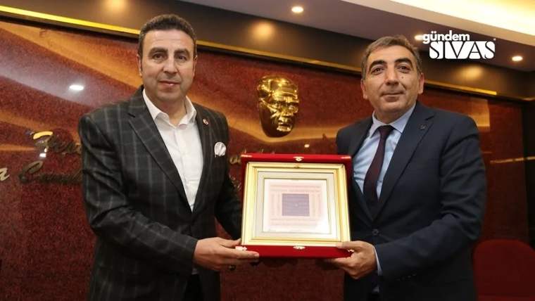 Bir kişi, diğerine plaket verirken gülümsüyor; arka planda bir heykel var.