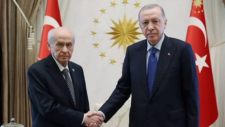 Takım elbiseli iki erkek, Türk bayrakları ve yıldız motifli arma önünde tokalaşıyor.