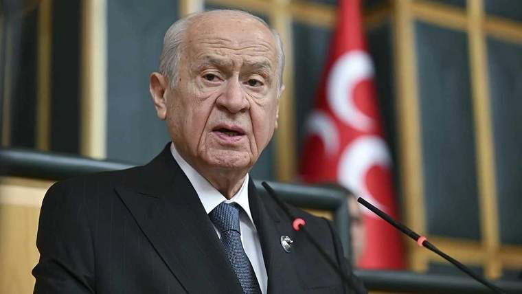 Kürsüde konuşan yaşlı bir erkek, koyu renk takım elbise ve kravatla mikrofonun önünde duruyor.