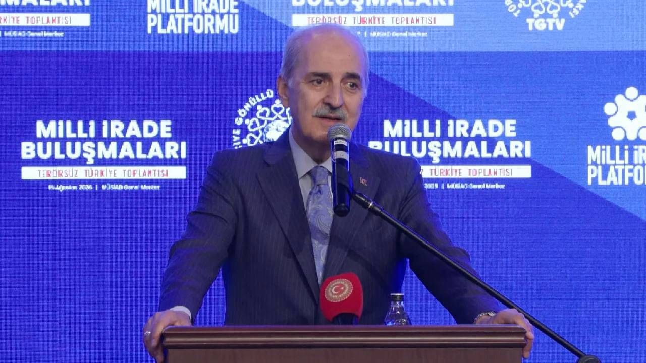 Kürsüde konuşan bir kişi, “Milli İrade Buluşmaları” yazılı mavi fonda mikrofon önünde duruyor.