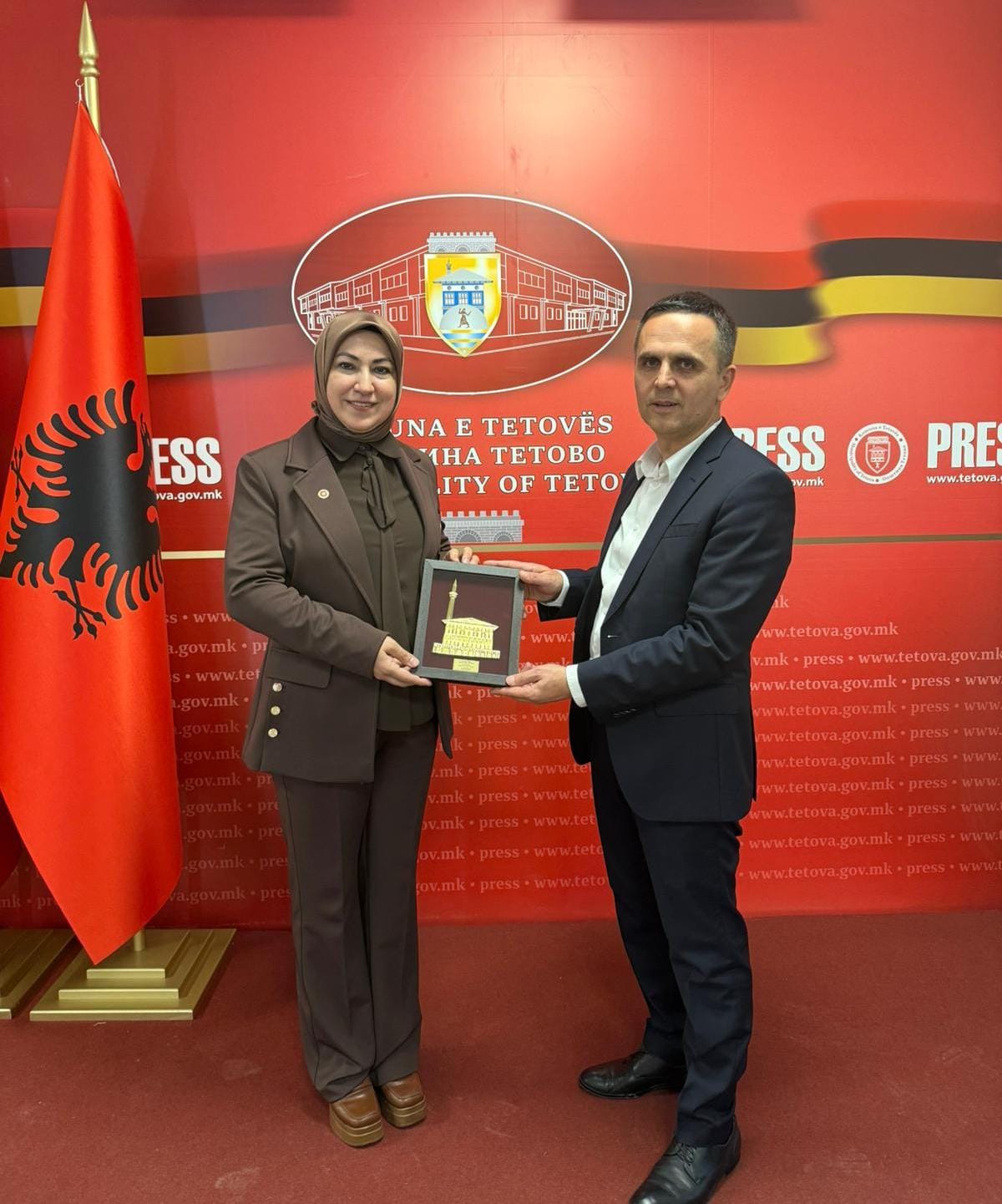 Kırmızı bayrak önünde bir kadın ve erkek, üzerinde bina resmi bulunan plaketi birlikte tutuyor.