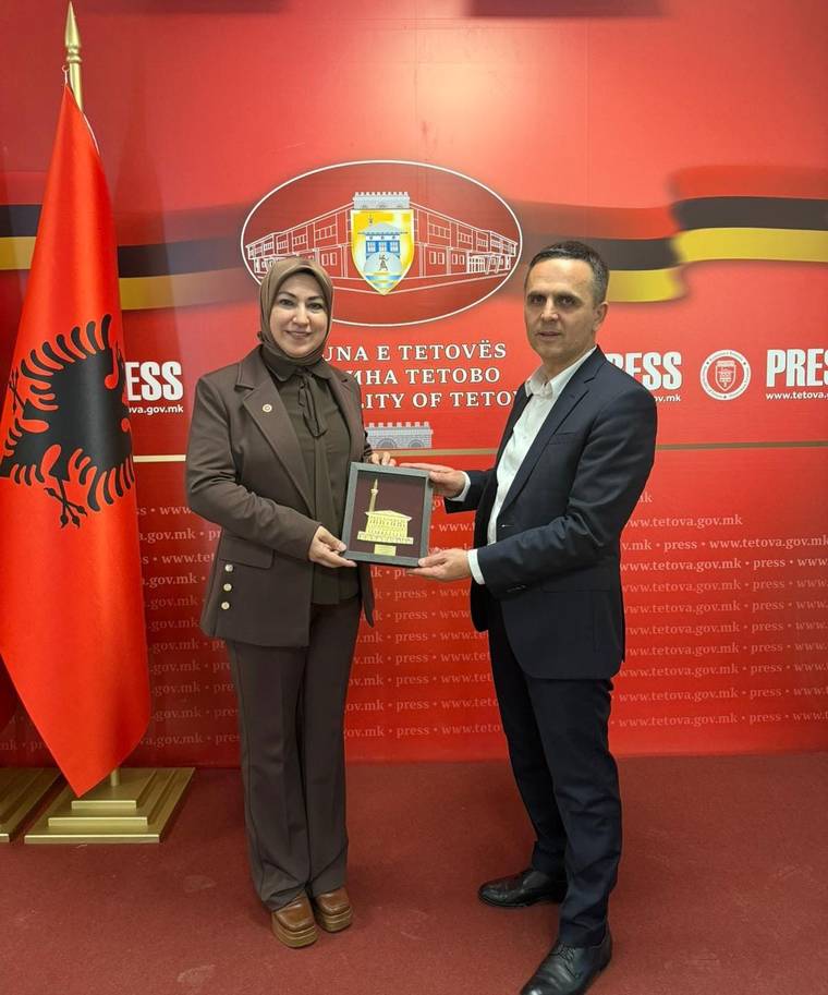Kırmızı bayrak önünde bir kadın ve erkek, üzerinde bina resmi bulunan plaketi birlikte tutuyor.