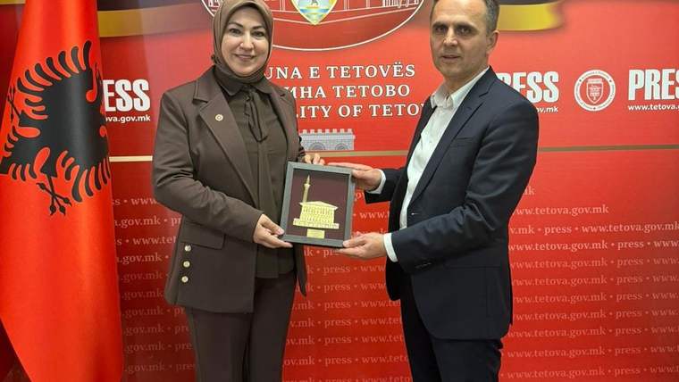 Kırmızı bayrak önünde bir kadın ve erkek, üzerinde bina resmi bulunan plaketi birlikte tutuyor.