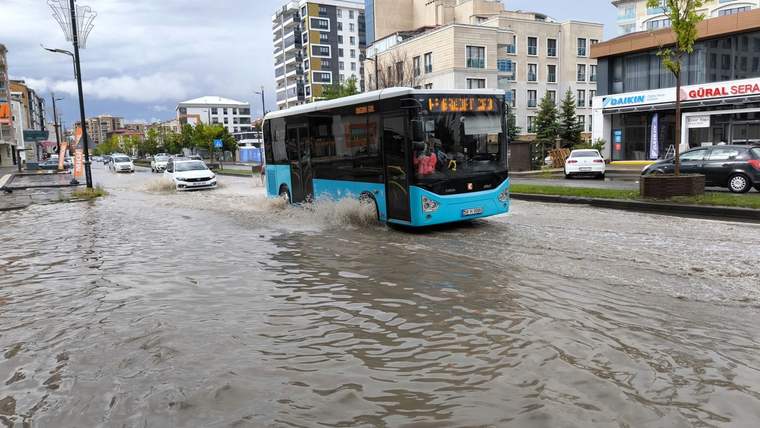 Su birikintileriyle kaplanan caddede turkuaz bir belediye otobüsü ilerlerken çevrede otomobiller ve binalar görülüyor.