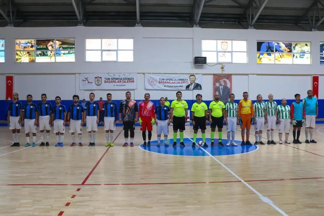 Kapalı spor salonundaki futsal sahasında, farklı renklerde formalar giyen sporcular ve hakemler yan yana duruyor.