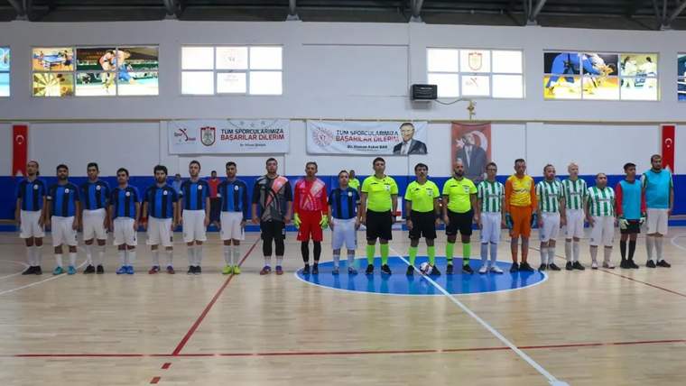 Kapalı spor salonundaki futsal sahasında, farklı renklerde formalar giyen sporcular ve hakemler yan yana duruyor.