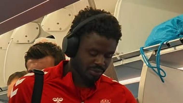 Uçak kabininde kırmızı spor kıyafetli iki kişi, koltukların arasında duran birinin telefonuna bakıyor.