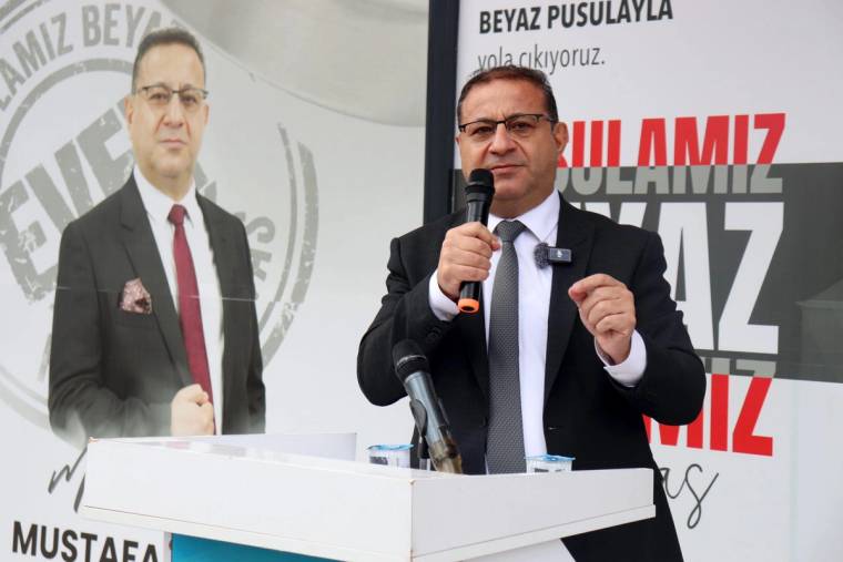 Mikrofonla konuşan bir kişi, üzerinde Mustafa Eken yazan afişin önünde kürsüde duruyor.