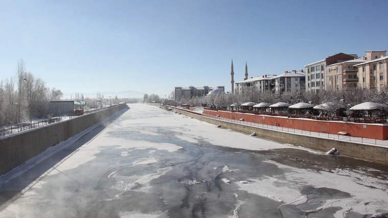 Buzla kaplı geniş bir nehir yatağı, kıyısındaki binalar, ağaçlar ve iki minareyle birlikte görülüyor.