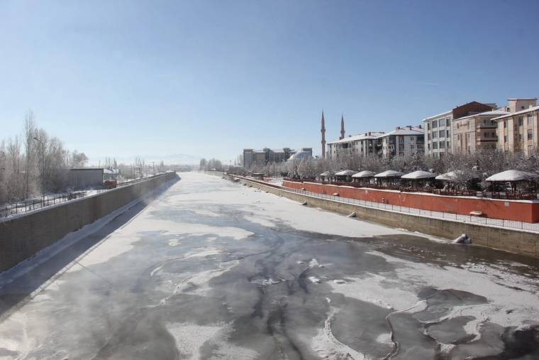 Buzla kaplı geniş bir nehir yatağı, kıyısındaki binalar, ağaçlar ve iki minareyle birlikte görülüyor.