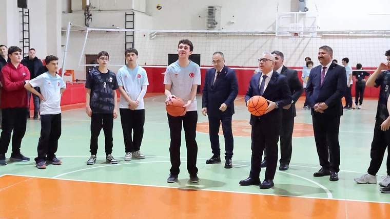 Basketbol topu tutan bir kişi, spor salonunda diğer öğrencilerle birlikte. Arka planda voleybol sahası görünmekte.