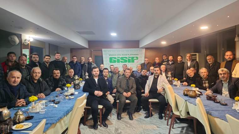 Toplantı salonunda masalar çevresinde oturan ve ayakta duran kalabalık bir grup, kamera karşısında poz veriyor.