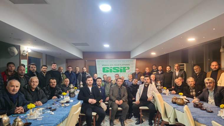 Kapalı salonda uzun masaların çevresinde oturan ve ayakta duran kalabalık grup, arkada GİSİP yazılı pankart