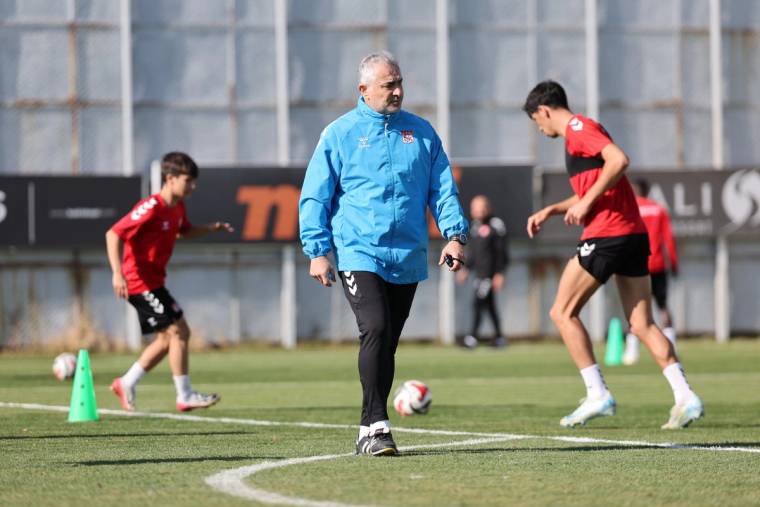 Mavi eşofmanlı bir kişi, çim sahada futbolcuların antrenmanı sırasında yürürken görülüyor.