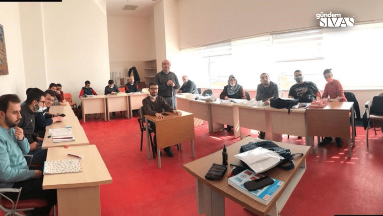 Kırmızı halılı sınıfta masalarda oturan yetişkinler ve ortada ayakta duran bir kişi görülüyor.