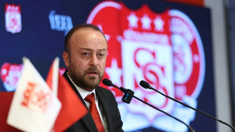 Takım armalı bir kürsü önünde takım elbiseli bir kişi, mikrofonlar arasında konuşuyor.