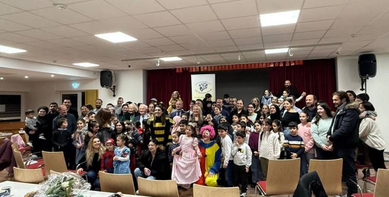 Perde önünde çocuklar ve yetişkinler birlikte poz verirken, ön sırada kostümlü çocuklar görülüyor.