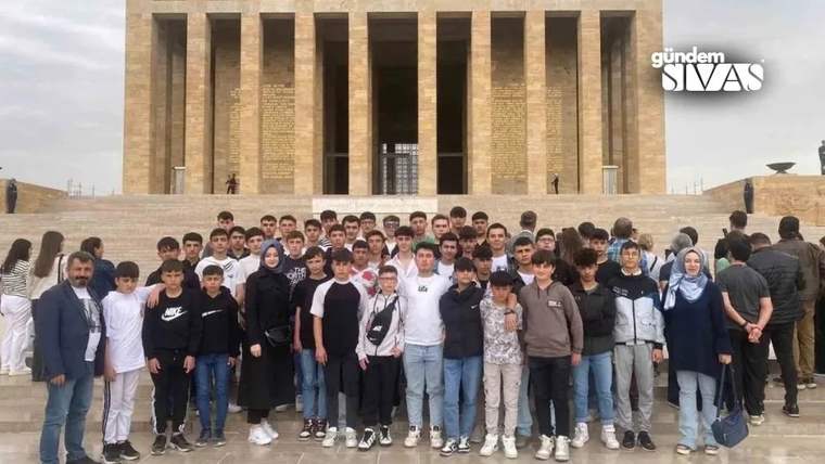 Büyük bir anıt yapısının önünde, çok sayıda öğrenci ve yetişkin toplu halde duruyor.