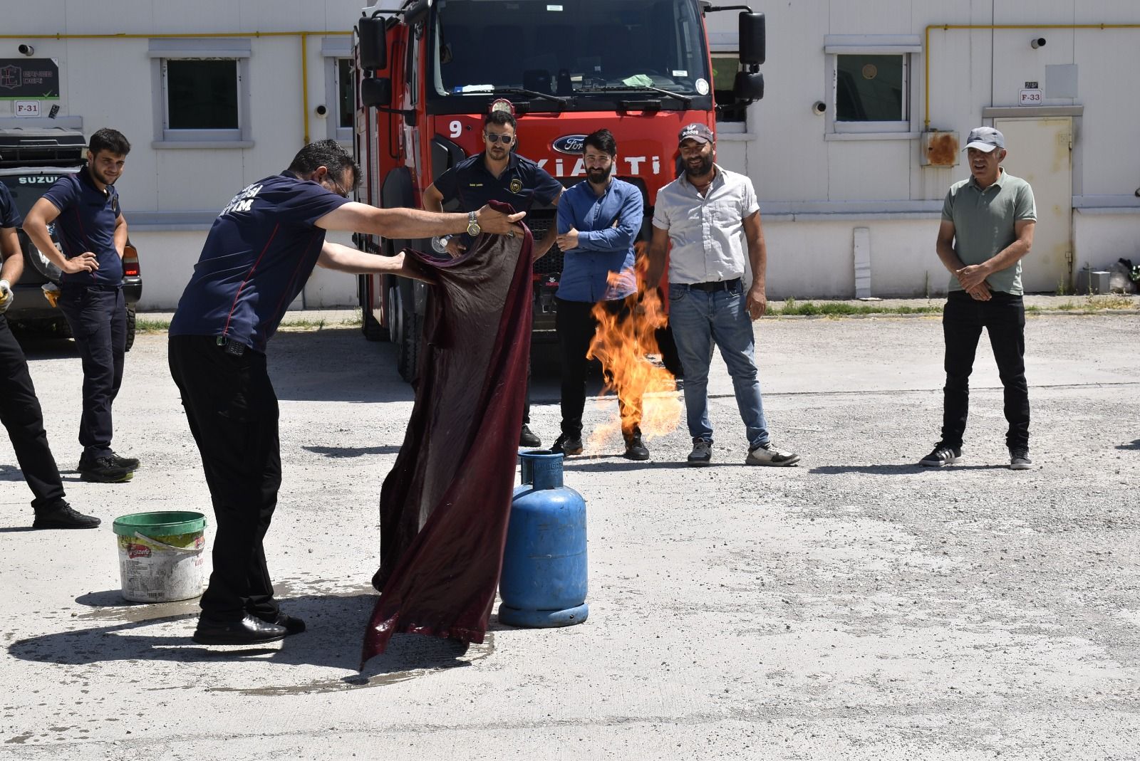 Beş kişinin önünde alev alan tüp bulunan alanda, bir kişi yangın battaniyesini tutuyor; itfaiye aracı arkada.