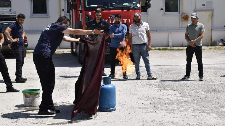 Beş kişinin önünde alev alan tüp bulunan alanda, bir kişi yangın battaniyesini tutuyor; itfaiye aracı arkada.