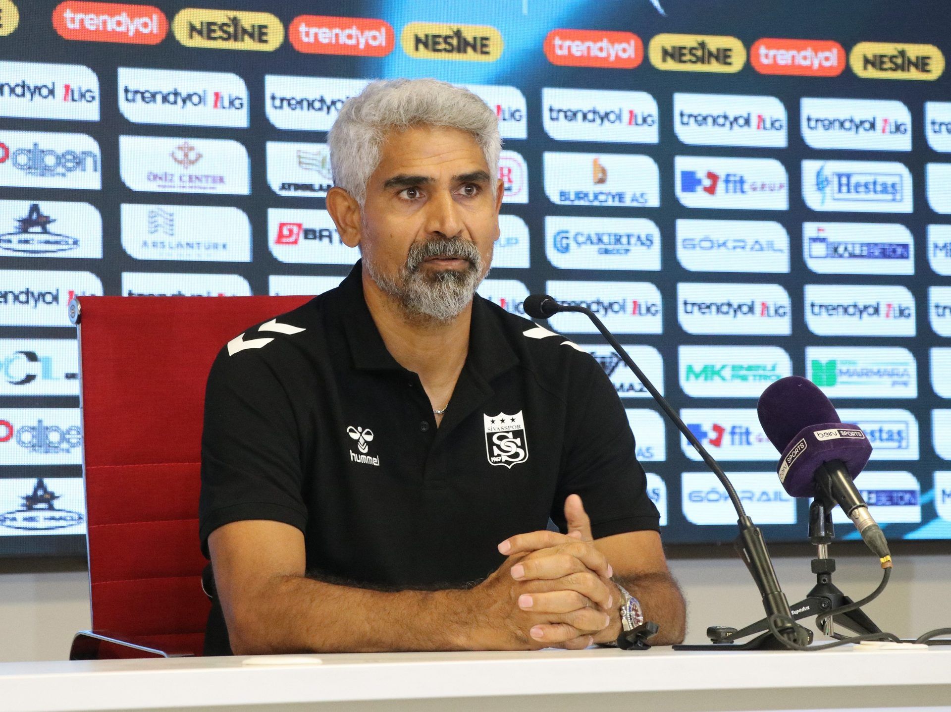 Spor giyim markalı polo tişört giyen bir kişi, sponsor panosu önünde mikrofonların yanında oturuyor.