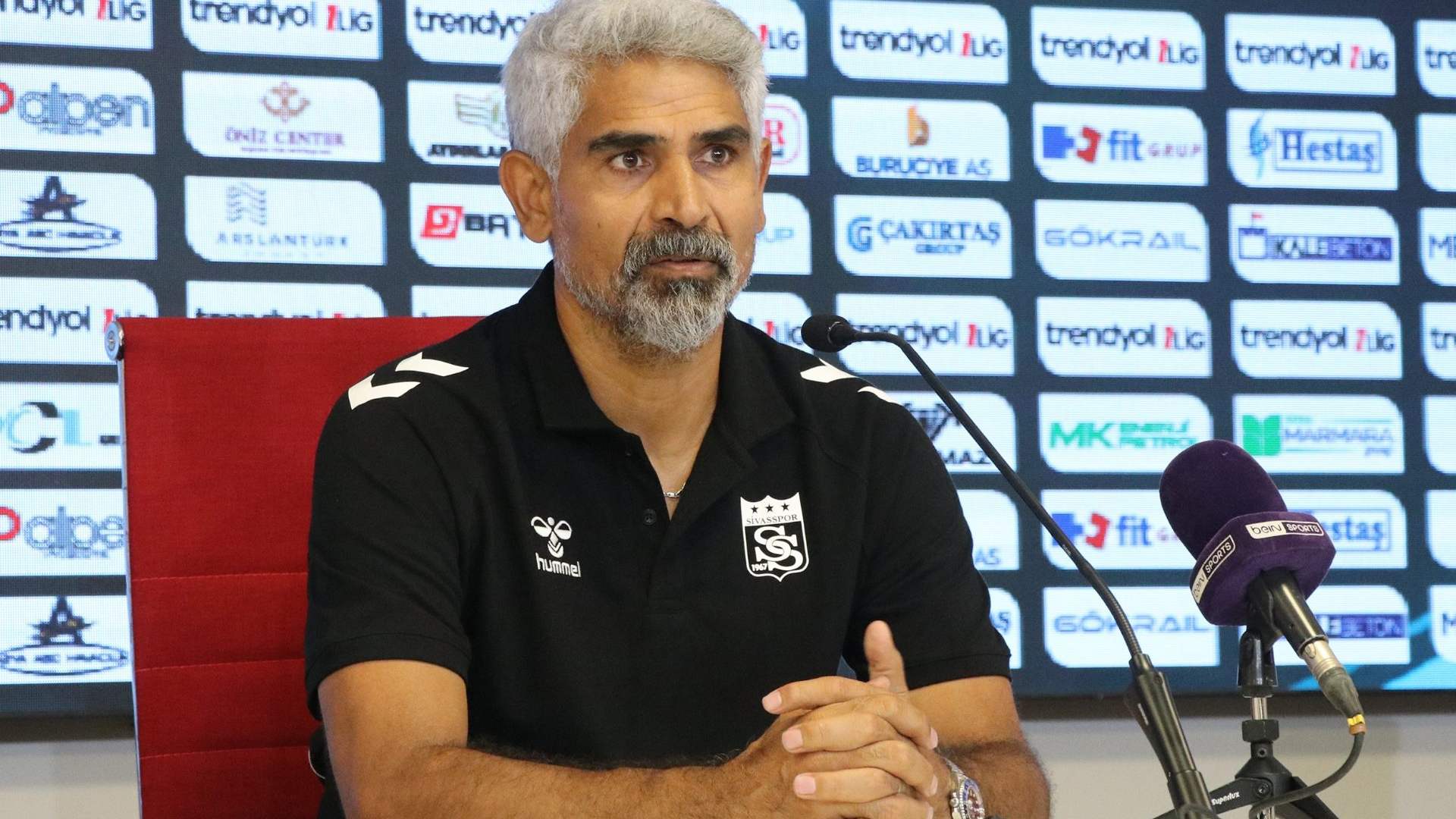 Spor giyim markalı polo tişört giyen bir kişi, sponsor panosu önünde mikrofonların yanında oturuyor.
