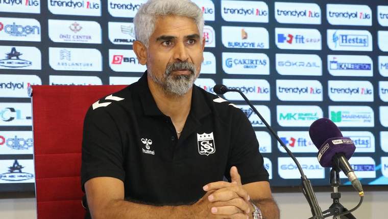 Spor giyim markalı polo tişört giyen bir kişi, sponsor panosu önünde mikrofonların yanında oturuyor.