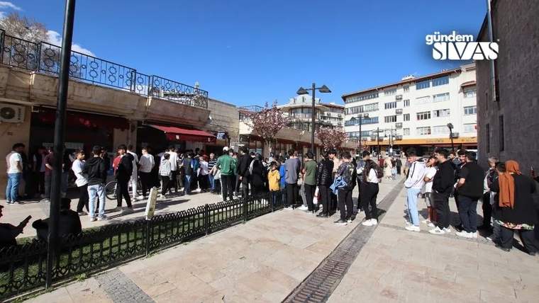 Bir binanın önünde, kaldırım boyunca uzanan sırada bekleyen kalabalık insanlar görülüyor.