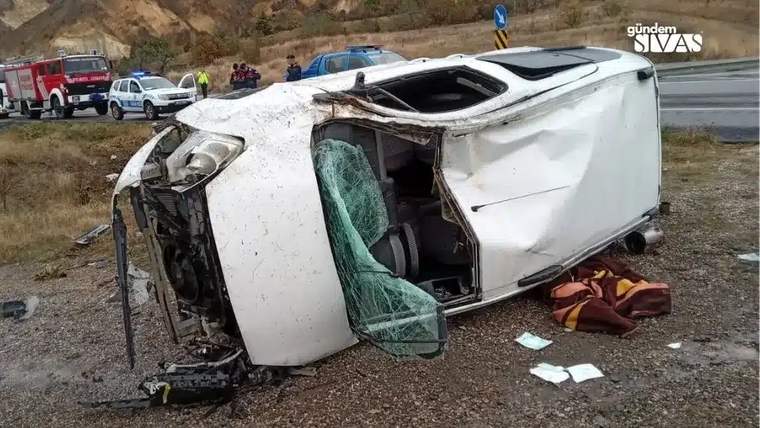 Yol kenarında yan yatmış beyaz otomobilin camları kırılmış; çevresinde itfaiye aracı ve başka araçlar bulunuyor.