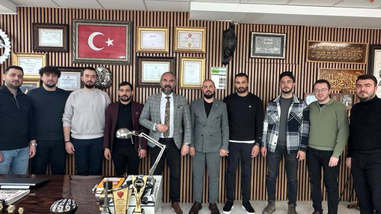 Masa önünde yan yana duran on kişi, arkalarında Türk bayrağı ve çok sayıda çerçeveli belge bulunan bir ofiste görülüyor.