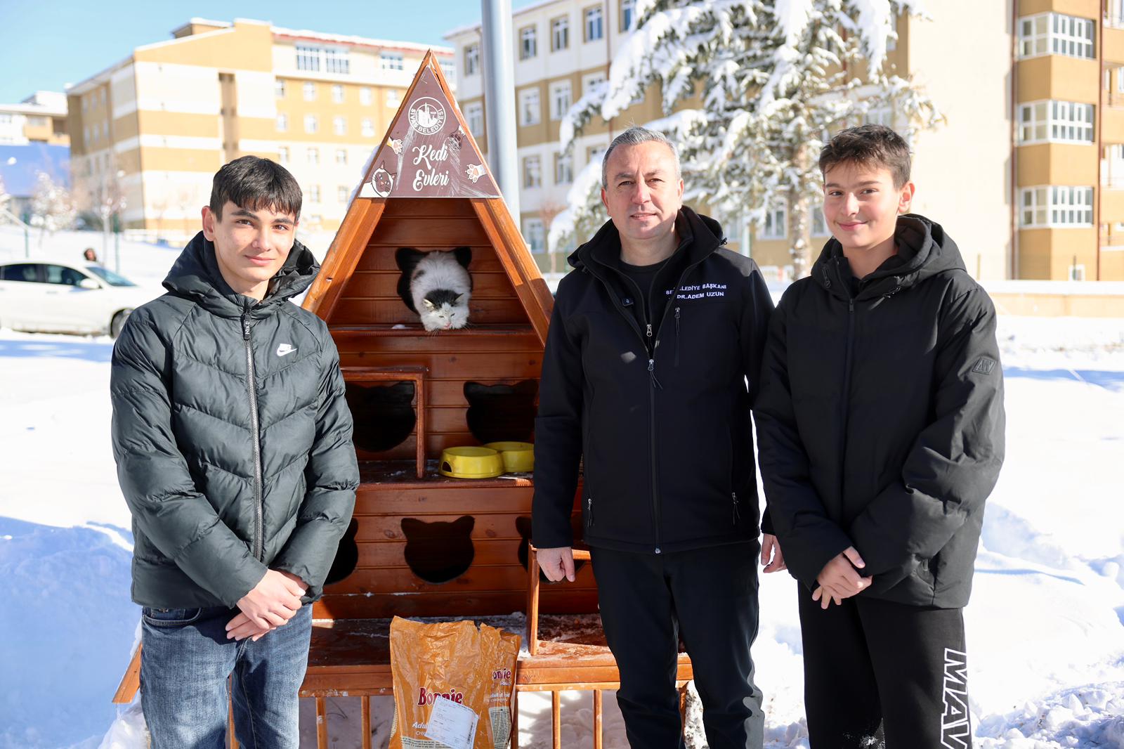 Kış şartlarında sokak kedileri için yapılmış bir kedi evi, yanında iki genç ve bir yetişkin duruyor.
