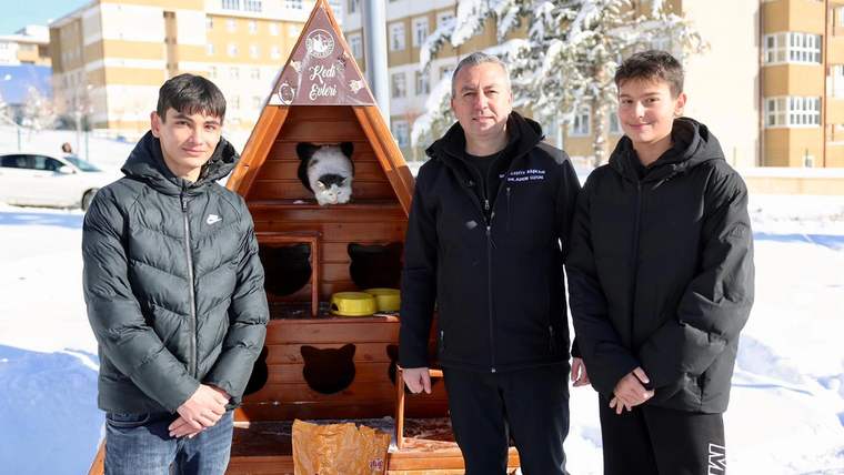 Kış şartlarında sokak kedileri için yapılmış bir kedi evi, yanında iki genç ve bir yetişkin duruyor.