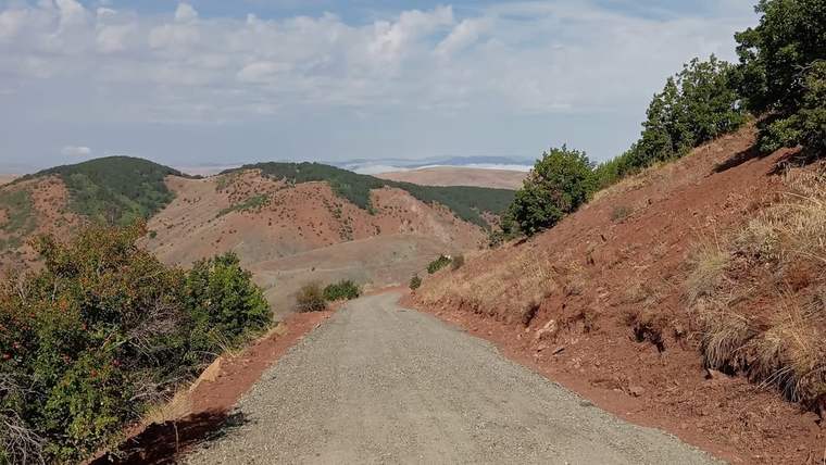Kırmızı toprak ve çakıl ile kaplı bir yol, dağlık bir alanda ilerliyor.