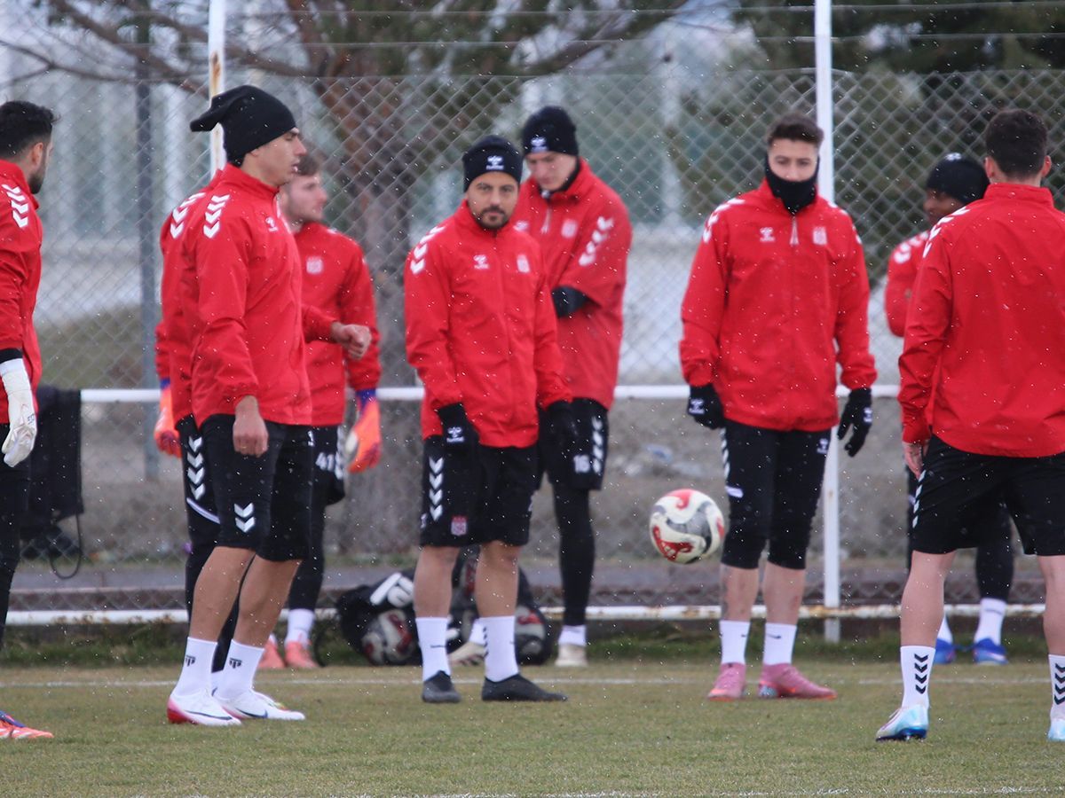 Kırmızı antrenman kıyafetleri giyen futbolcular, yağış altında sahada futbol toplarıyla bekliyor.