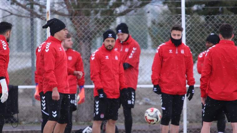 Kırmızı antrenman kıyafetleri giyen futbolcular, yağış altında sahada futbol toplarıyla bekliyor.