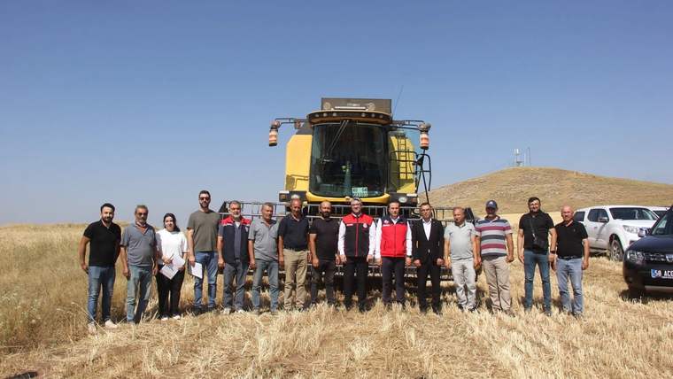 Hasat edilmiş tarlada, biçerdöverin önünde duran kalabalık bir grup ve çevrede park etmiş araçlar görülüyor.