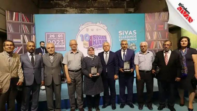 Kitap rafları önünde sekiz kişi, üzerinde “10. Sivas Kitap Günleri” yazan pano önünde yan yana duruyor.