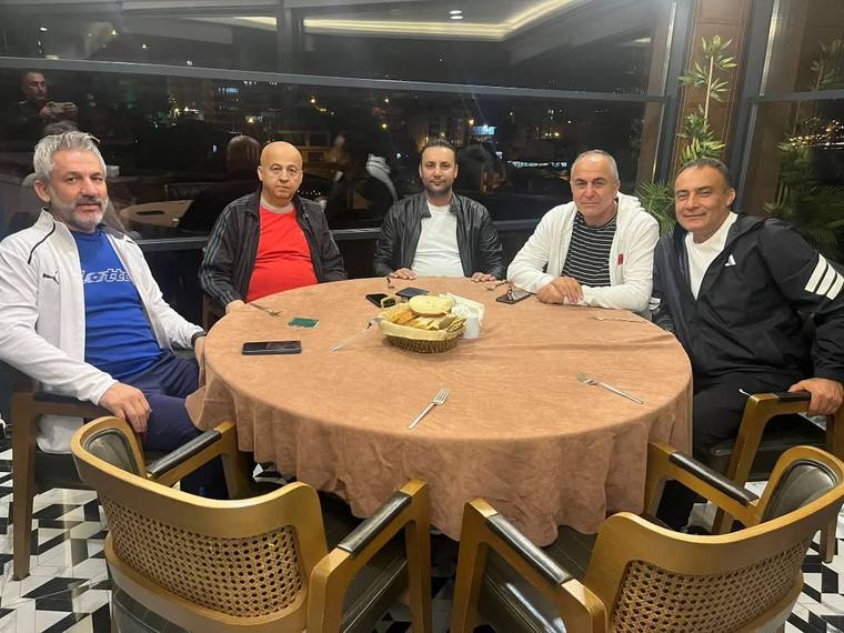 Restoran masasının etrafında oturan beş kişi, gece aydınlatılmış camların önünde poz veriyor.