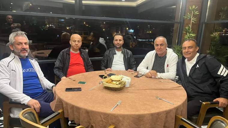 Restoran masasının etrafında oturan beş kişi, gece aydınlatılmış camların önünde poz veriyor.