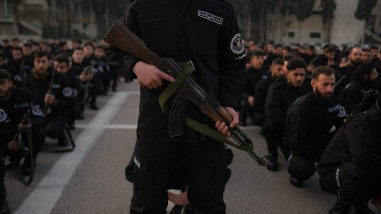 Siyah üniformalı, tüfek taşıyan bir kişi ön planda dururken arkasında çok sayıda kişi diz çöküyor.