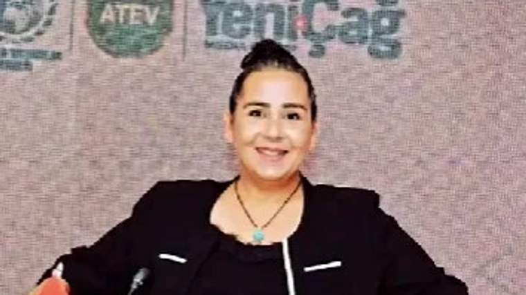 Kürsüde oturan bir kadın, elinde Türk bayrağıyla kameraya bakıyor; arka planda etkinlik logoları bulunuyor.