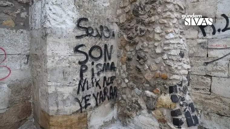Taş duvarın sıvalı bölümünde siyah sprey boyayla yazılmış yazılar ve çeşitli karalamalar görülüyor.